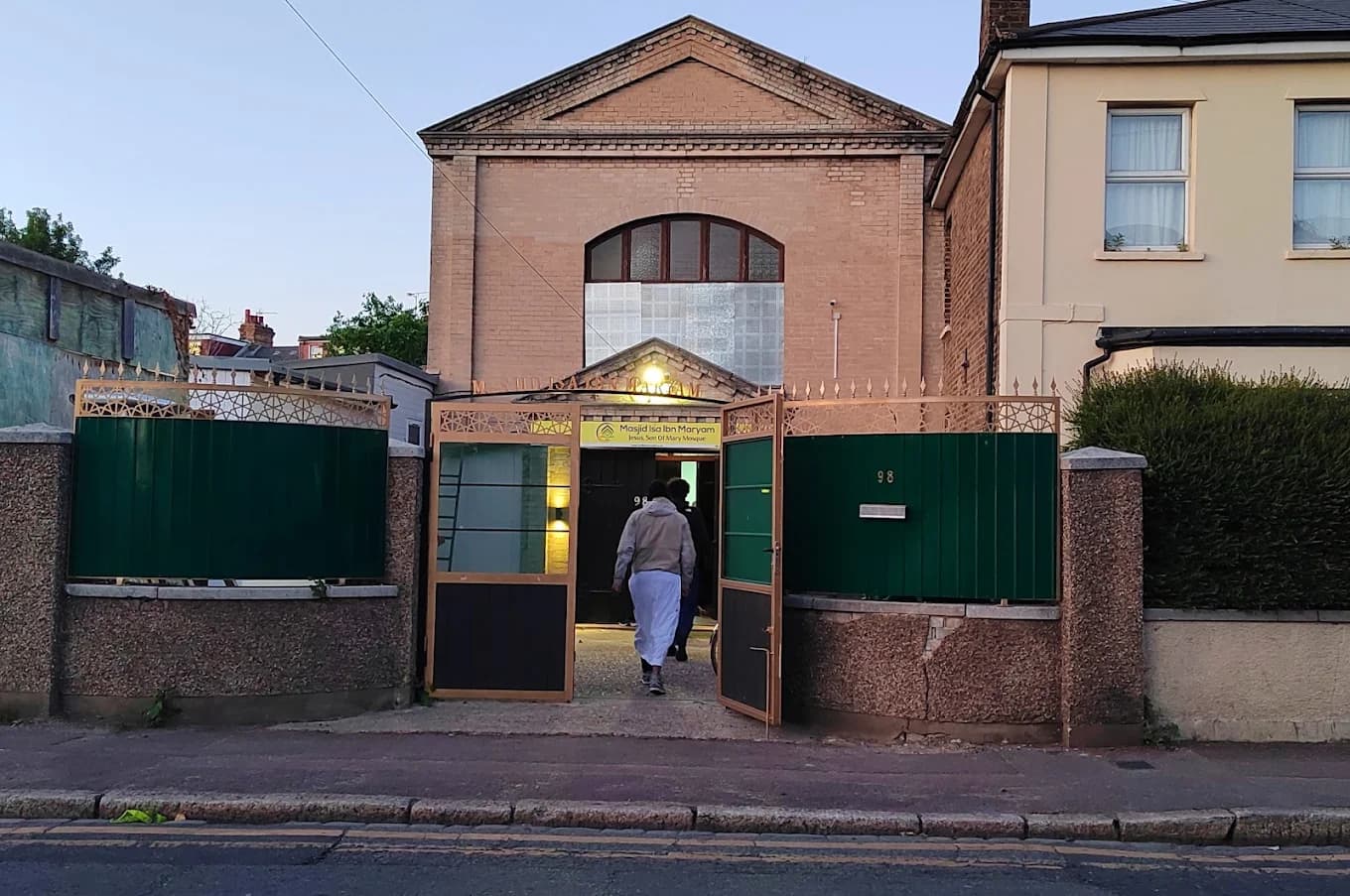 The masjid seen from across Dames Road at dusk, with green railings, gold gates open and worshippers walking in