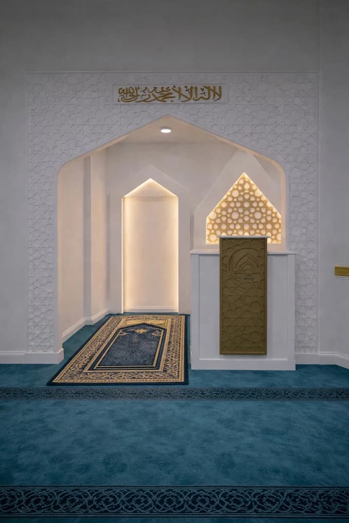 The mihrab: a white arched niche with carved geometric plasterwork and gold Arabic calligraphy above, a gold minbar panel beside it, and a patterned prayer rug on deep teal carpet